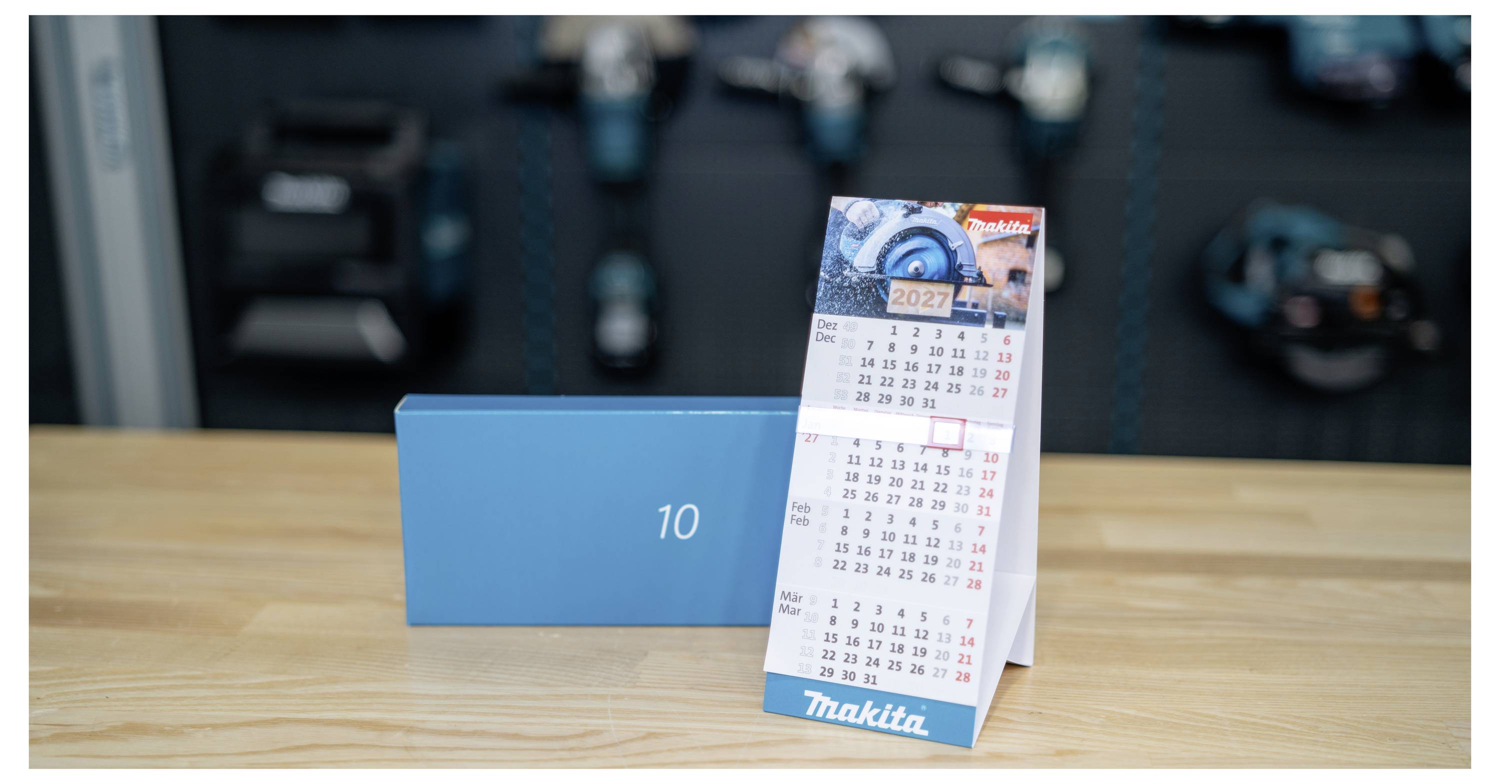Ein Makita-Schreibtischkalender steht auf einem Holztisch neben einer blauen Box mit der Aufschrift „10