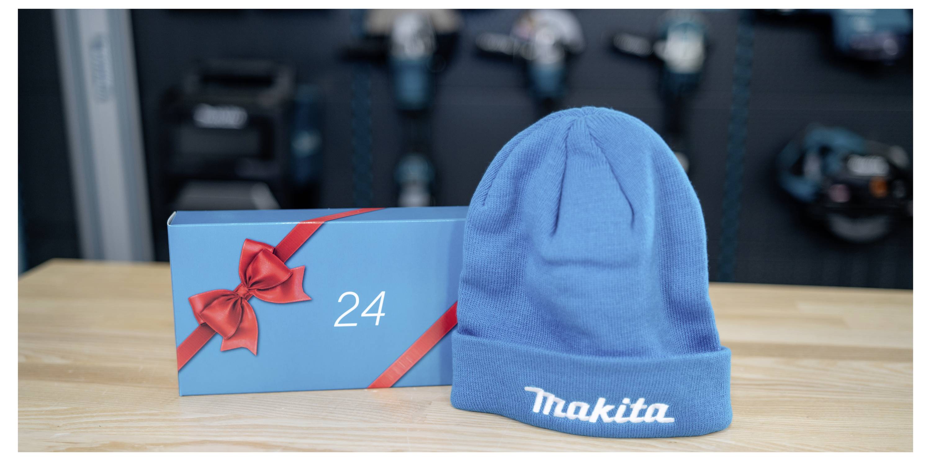 Blaue Makita-Beanie-Mütze neben einer blauen Geschenkbox mit rotem Band und der Nummer 24 auf einem Holztisch, mit unscharfen Maschinen im Hintergrund