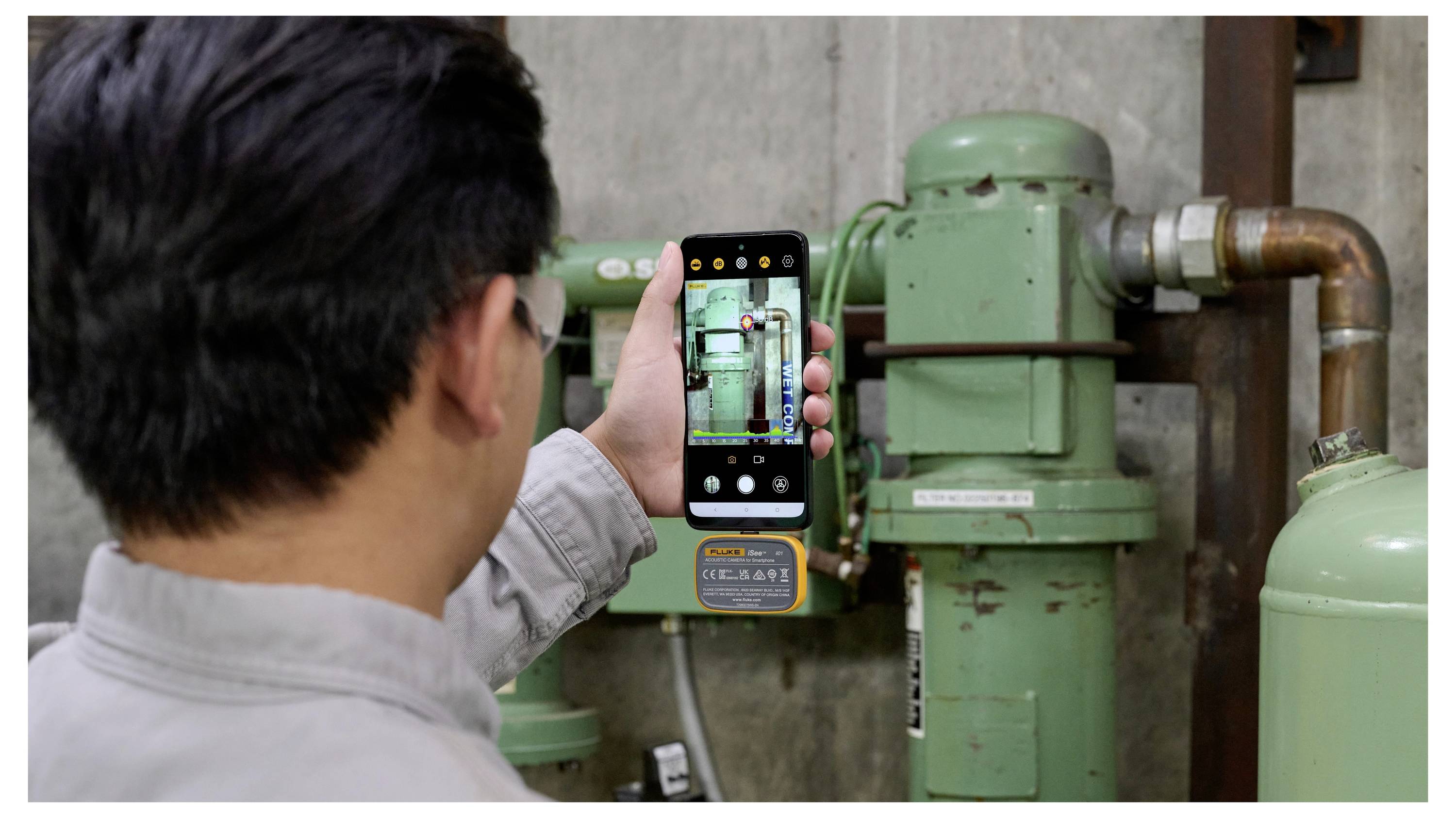 Person fotografiert grüne Industrierohre und Ausrüstung mit einem Smartphone und inspiziert Maschinen in einer Anlage.