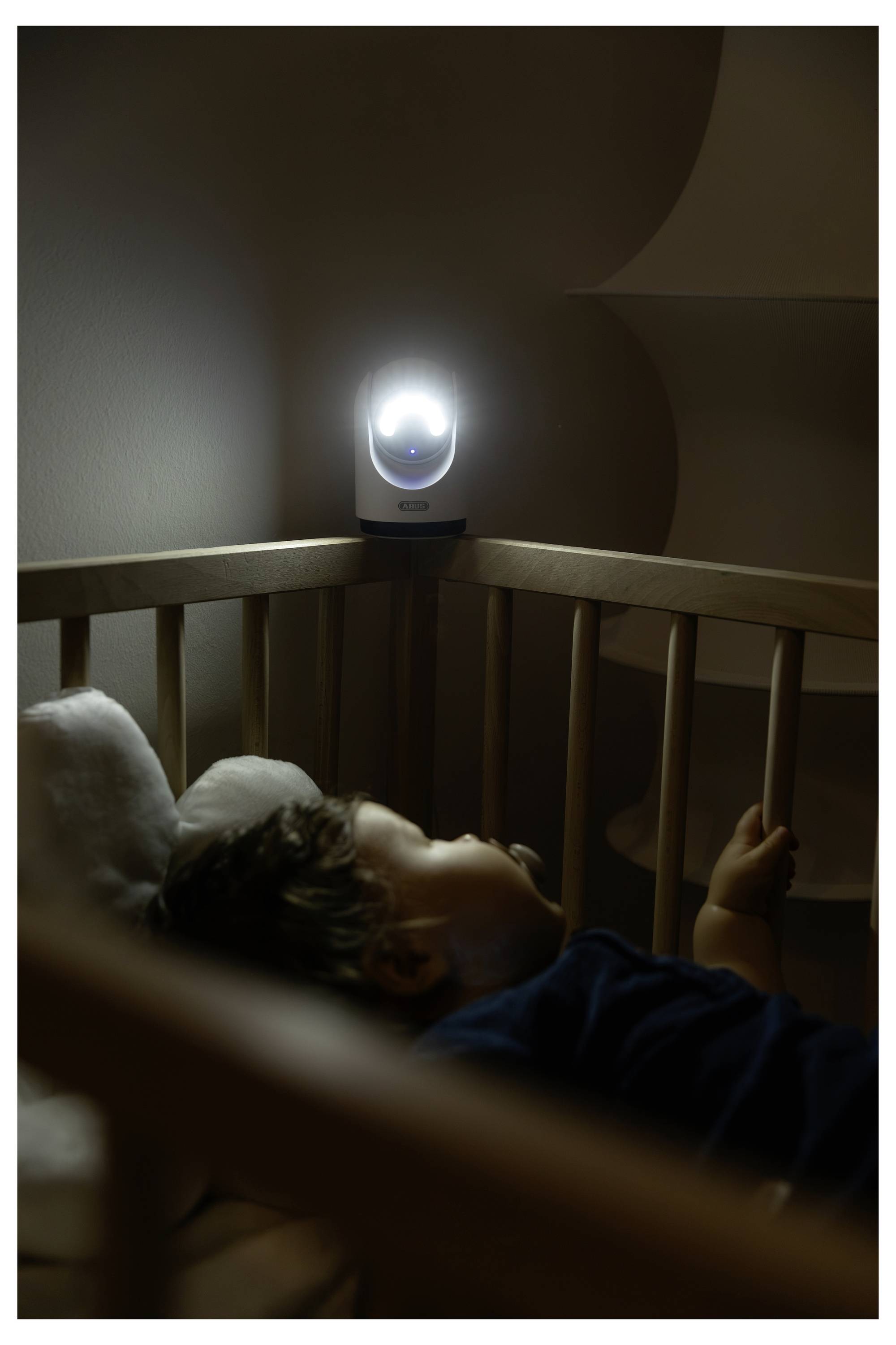 Baby schläft in einem Gitterbett, während ein leuchtendes Babyphone die Kinderstube in der Dunkelheit überwacht.