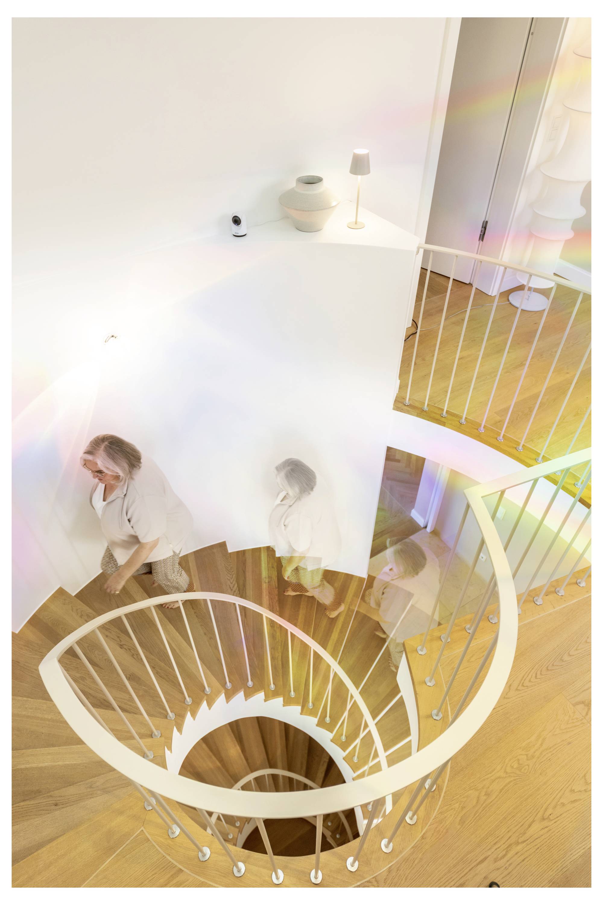 Ansicht von oben auf eine geschwungene weiße Treppe mit einer Person, die hinabsteigt; warme Holzböden und helle Inneneinrichtung sind sichtbar.