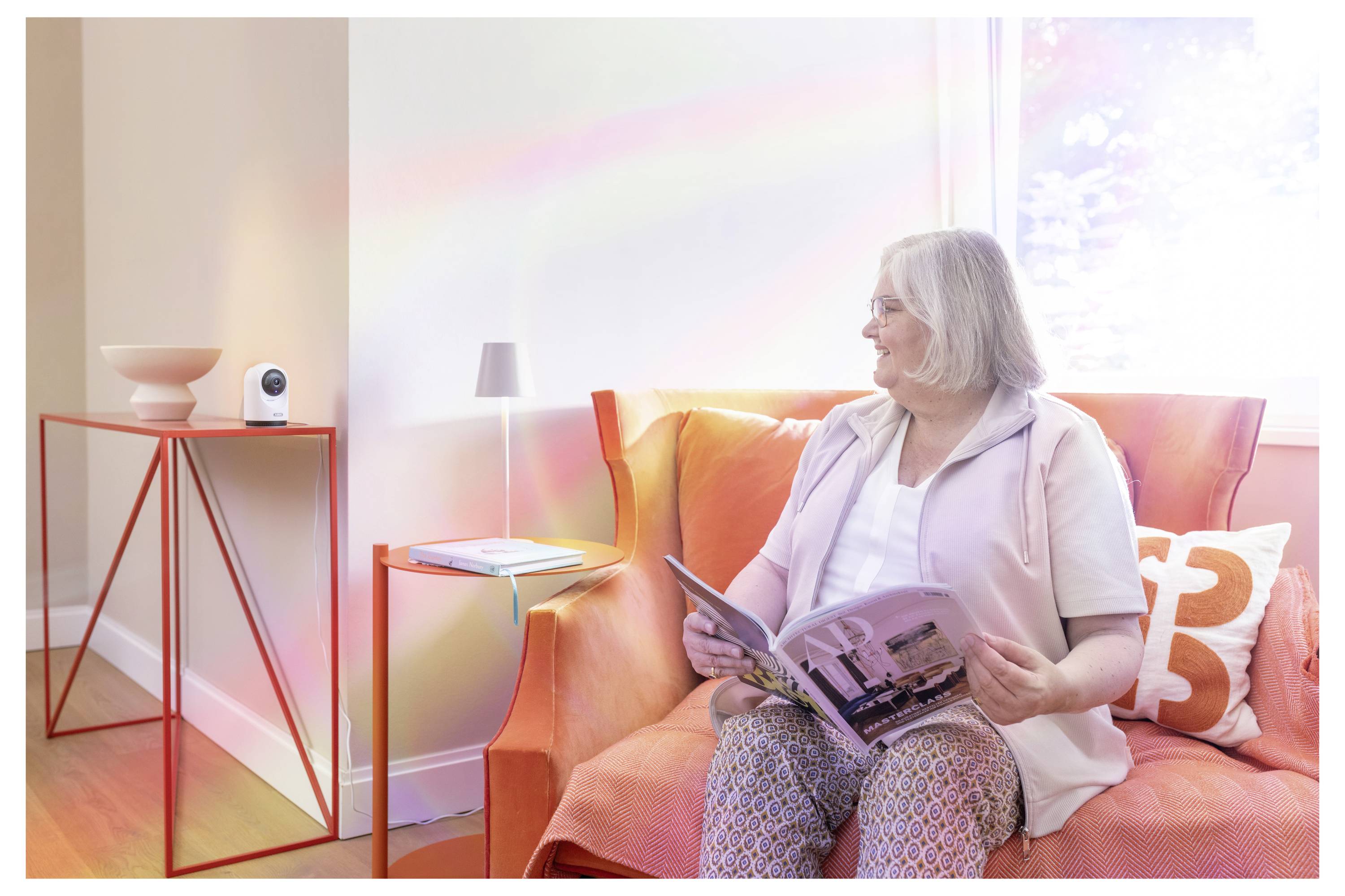 Eine ältere Frau sitzt auf einem orangefarbenen Sofa und liest eine Zeitschrift in einem hellen Wohnzimmer mit einem kleinen Beistelltisch und einer Lampe in der Nähe.
