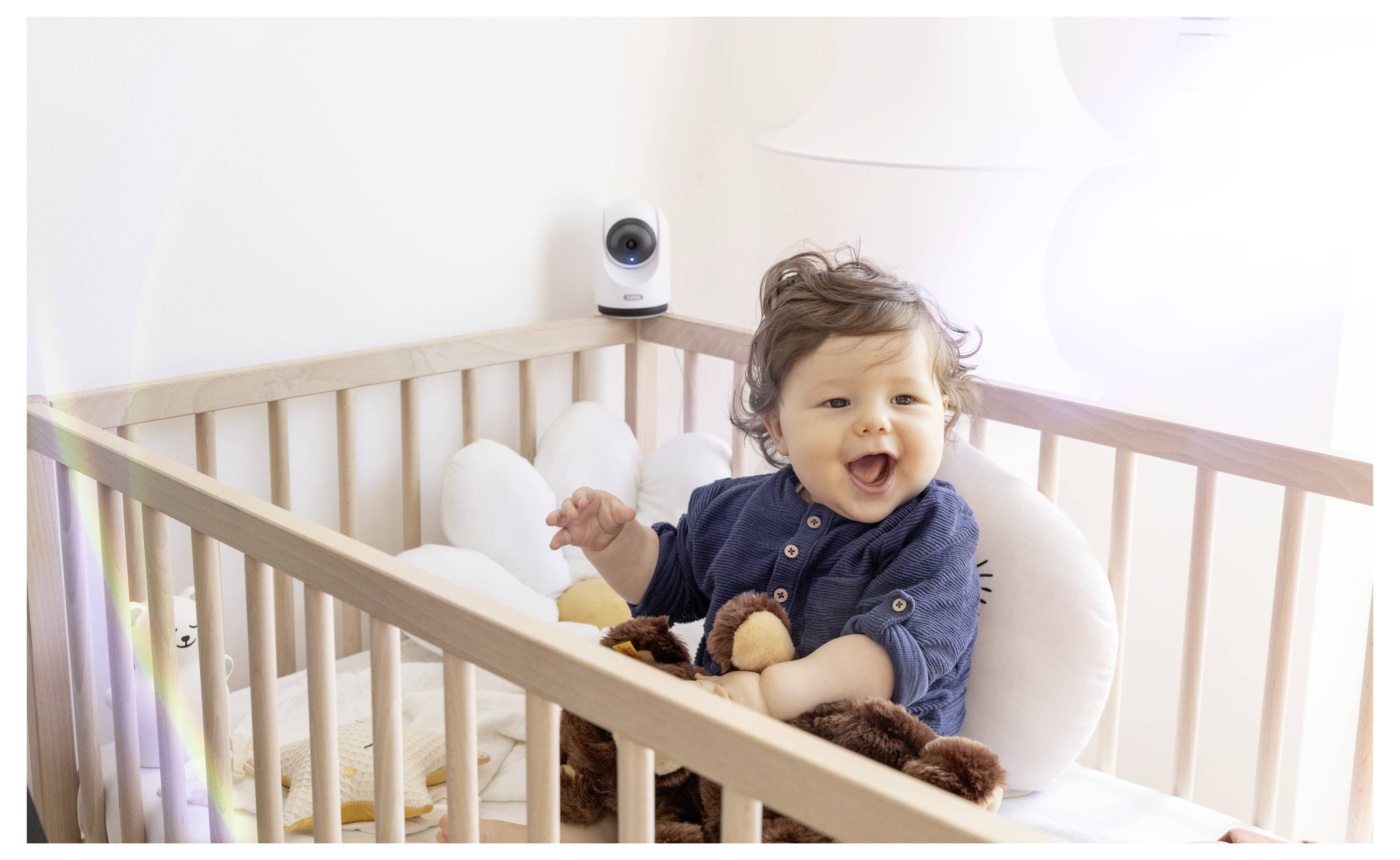 Lächelndes Baby sitzt in einem Holzgitterbett mit Plüschtieren und einem Babyphone in der Ecke.