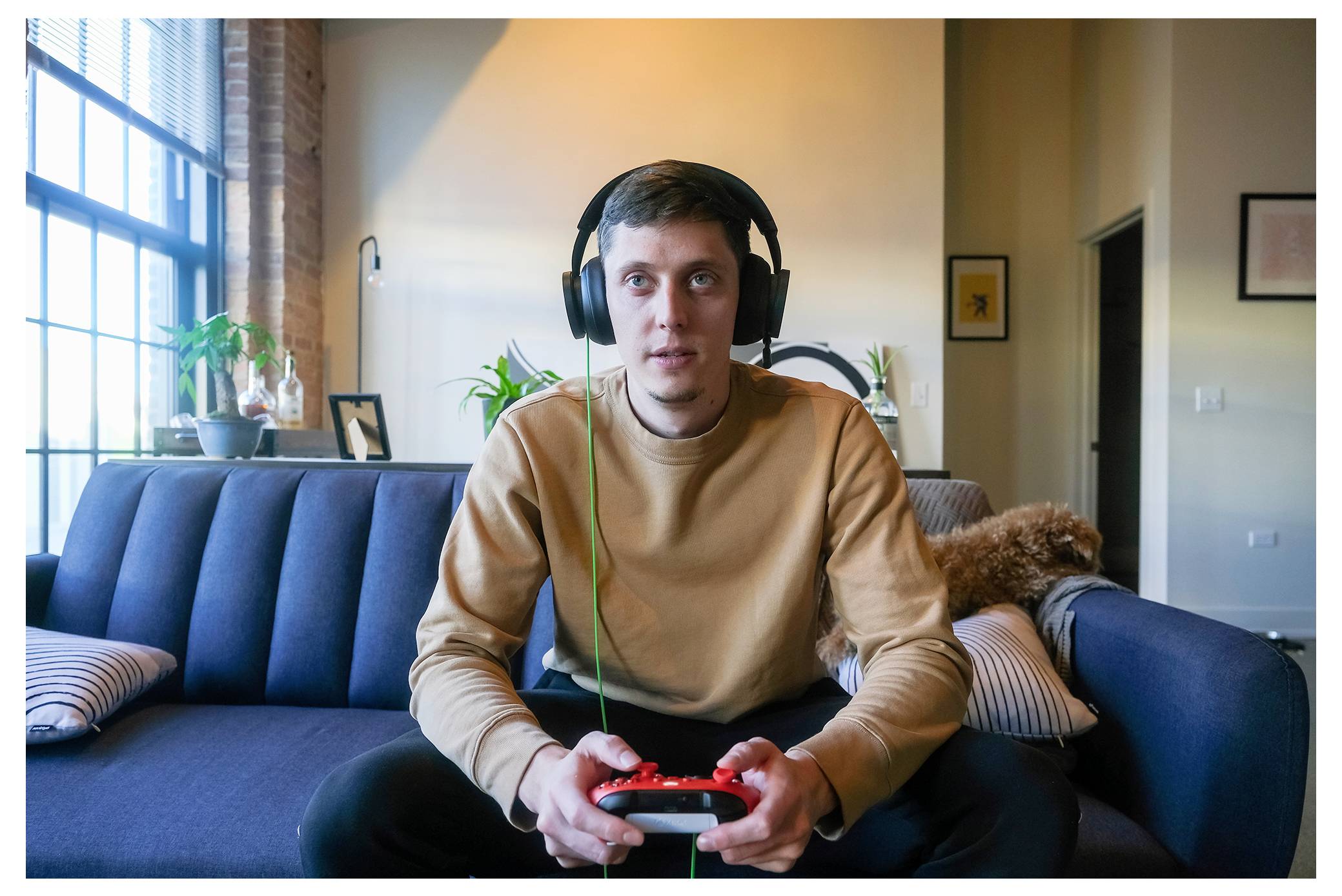 Junger Mann mit Kopfhörern spielt ein Videospiel auf einer Couch im Wohnzimmer und hält einen Controller in der Hand.