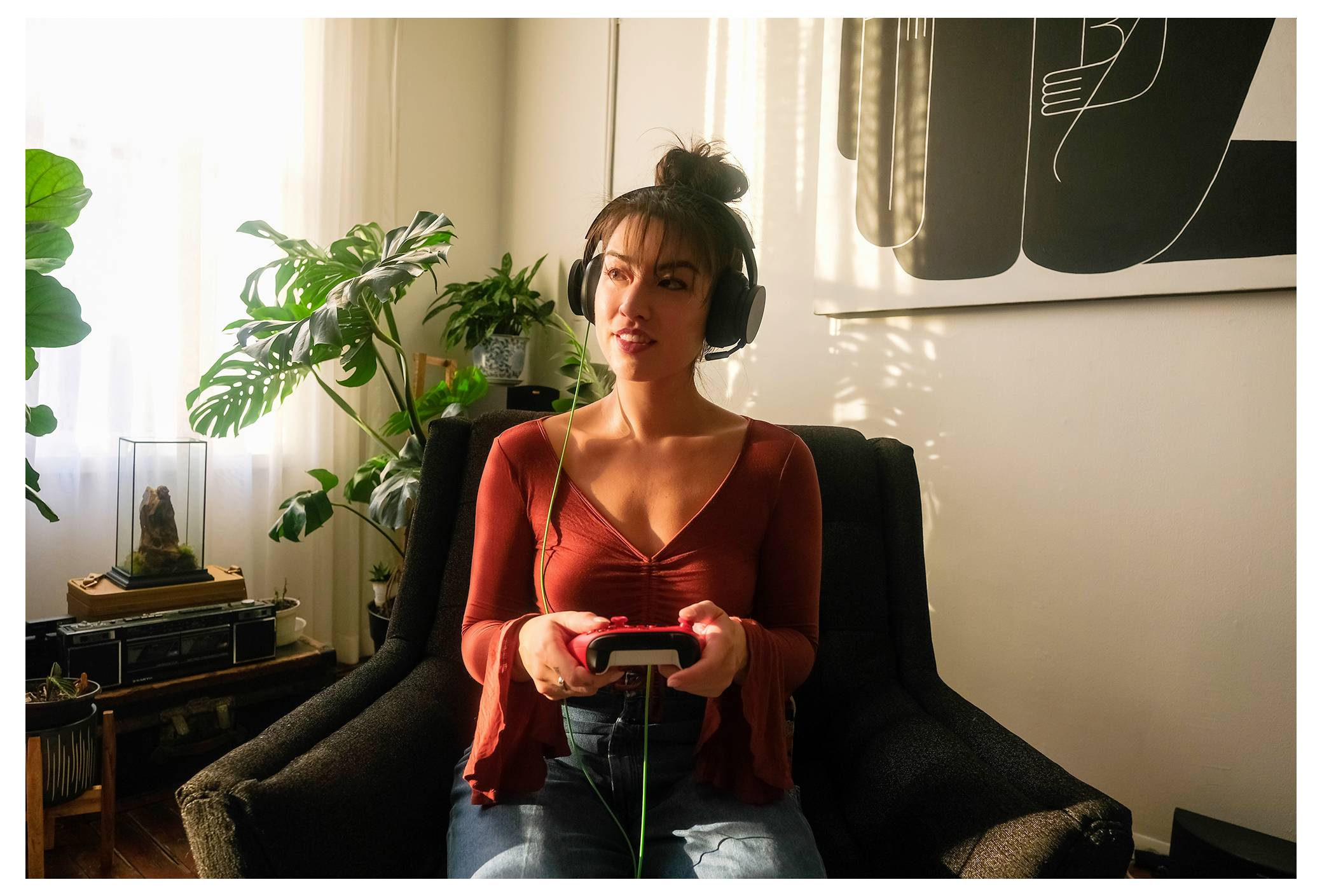 Frau mit Kopfhörern spielt ein Videospiel, während sie in einem Stuhl in einem sonnendurchfluteten Wohnzimmer sitzt.