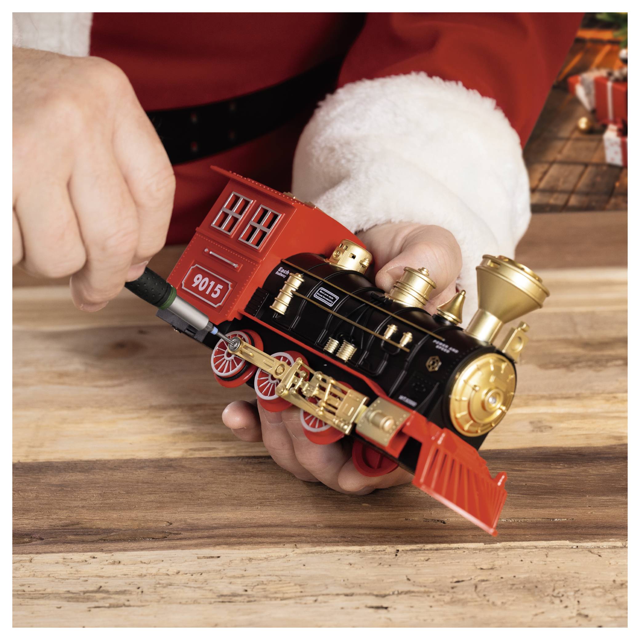 Hände halten eine rot-schwarze Spielzeug-Dampflokomotive auf einem Holztisch, mit einem kleinen Etikett 
