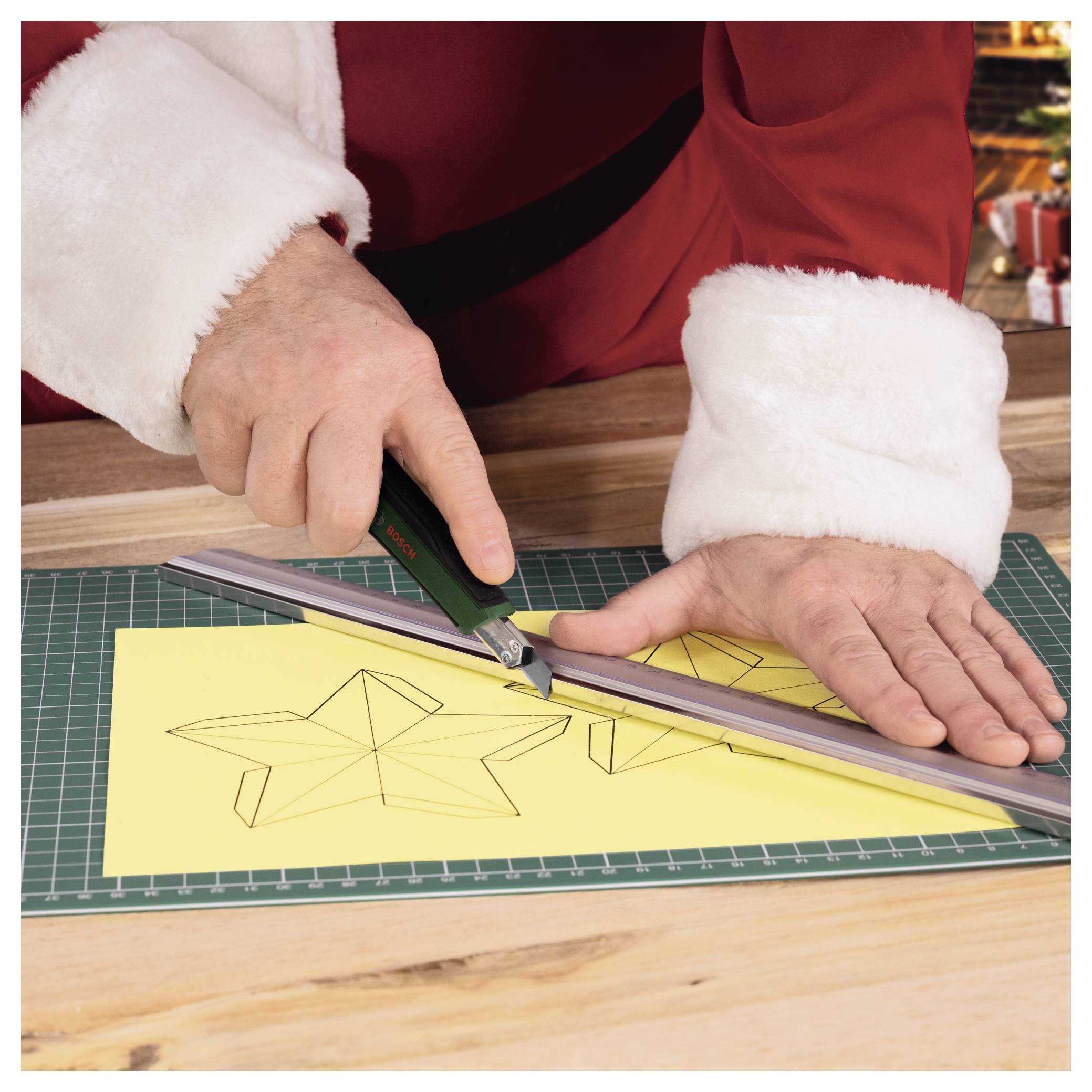 Der Weihnachtsmann in einem roten Anzug benutzt ein Lineal und ein Bastelmesser, um eine Sternform aus gelbem Papier auf einer Schneidematte auszuschneiden.