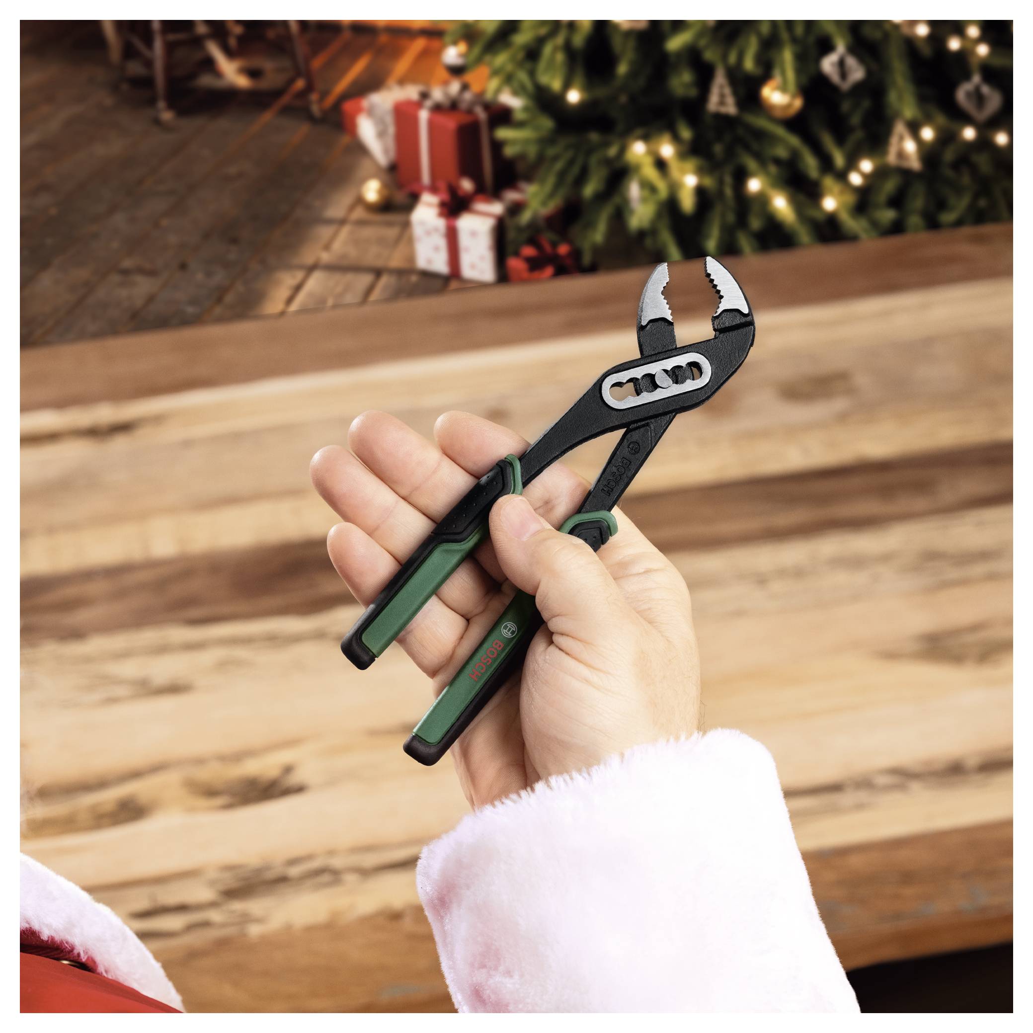 Eine mit Weihnachtsmannkleidung bekleidete Hand hält eine verstellbare Zange über einen Holztisch, mit einem geschmückten Weihnachtsbaum und eingepackten Geschenken im Hintergrund.