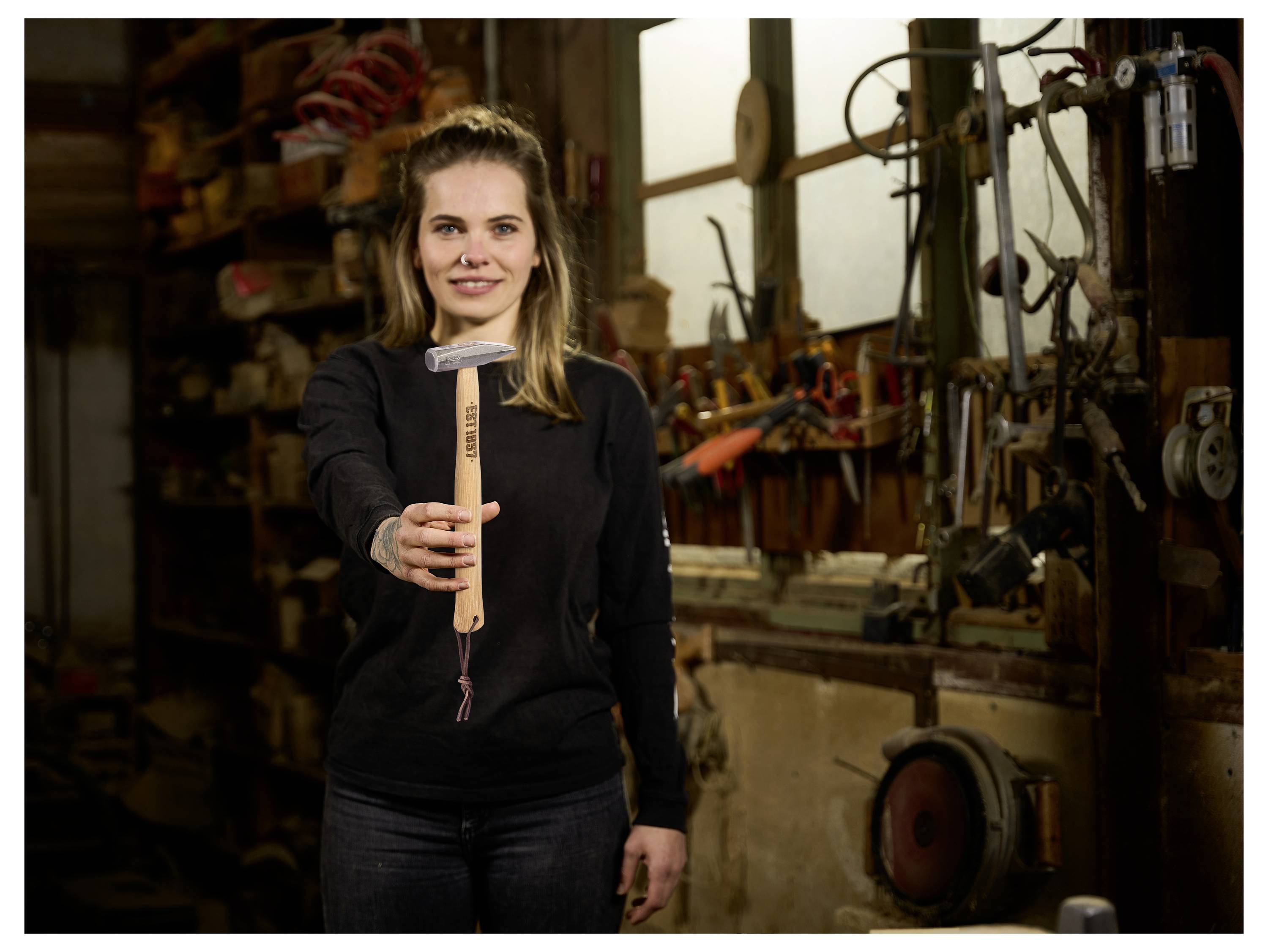 Frau in einer Werkstatt, die einen Hammer hochhält, steht vor Werkzeugen und Maschinen und deutet auf eine Handwerks- oder Reparaturumgebung hin.