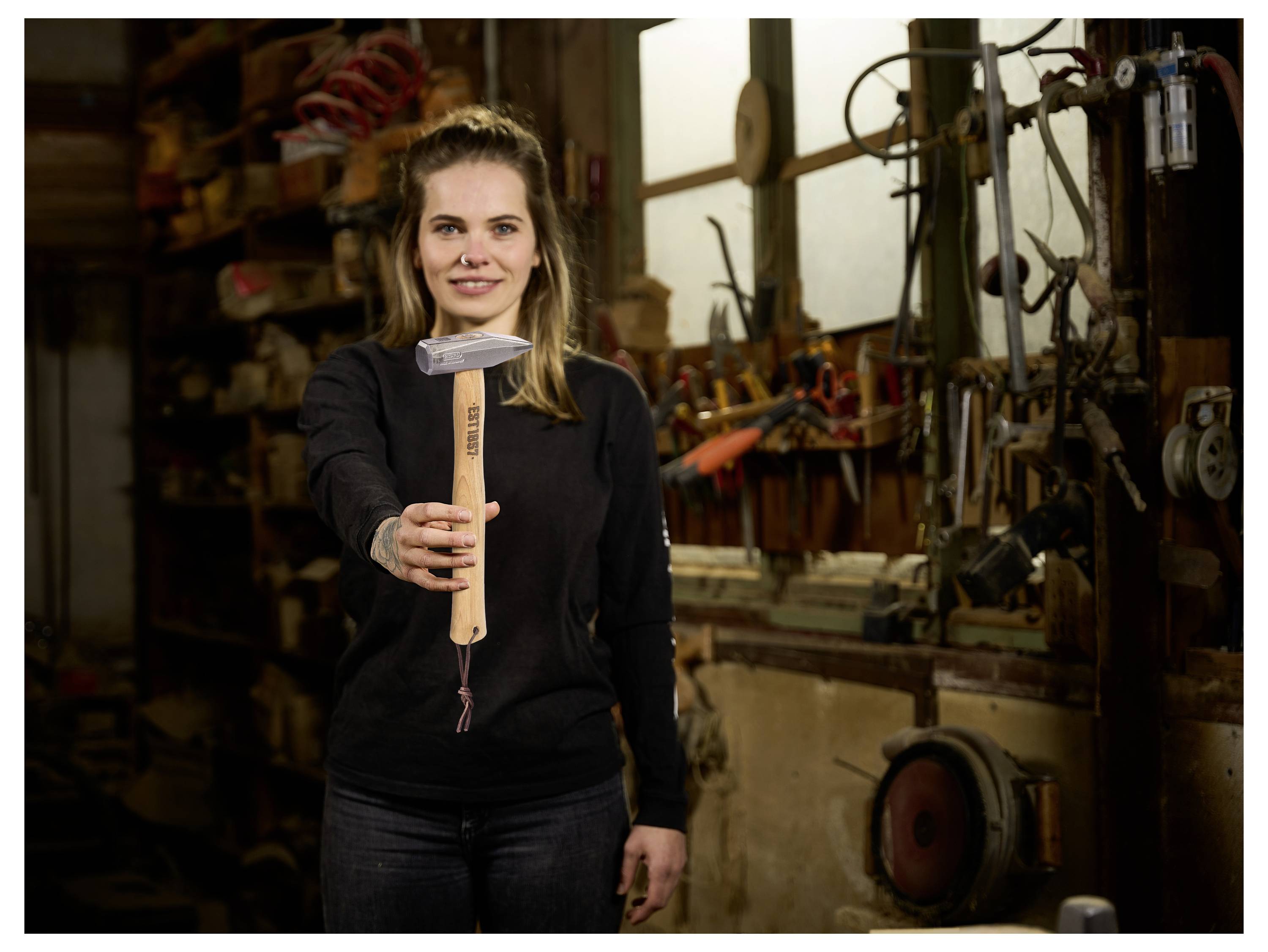 Frau in einer Werkstatt, die einen Hammer in Richtung Kamera hochhält, mit Werkzeugen und Maschinen im Hintergrund.