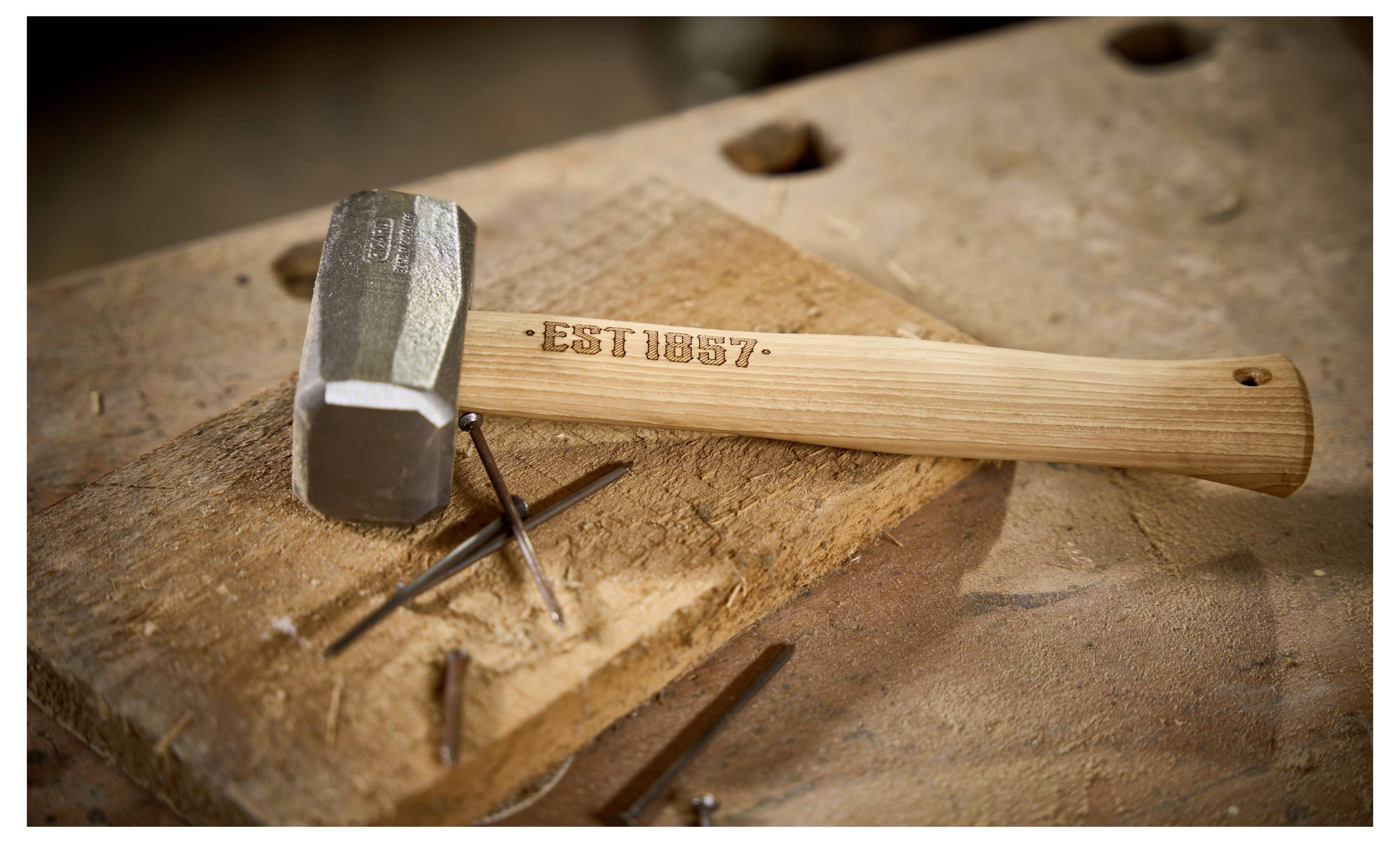 Ein Metallhammer mit Holzgriff ruht auf einer Werkbank neben kleinen Nägeln, wobei der Griff mit „EST 1957