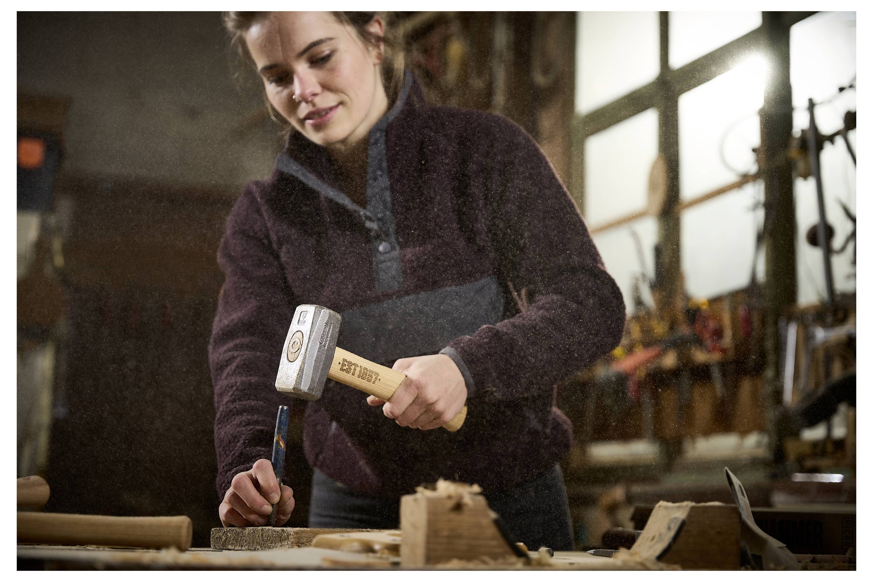Frau in einer Werkstatt, die einen Hammer und Meißel auf ein Holzstück an einer Werkbank verwendet.