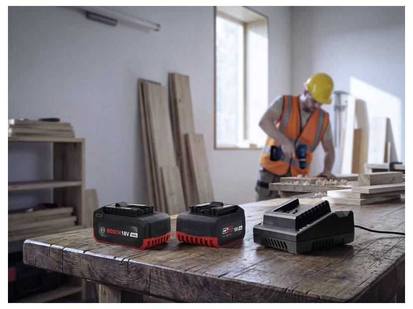 Baustellen-Foto eines Bauarbeiters mit Schutzhelm und Warnweste, der ein Elektrowerkzeug in einer Werkstatt verwendet, mit Batterien auf einer Werkbank.