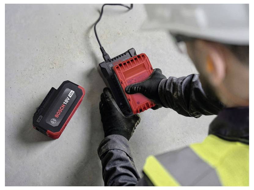 Arbeiter in Schutzausrüstung steckt ein rotes Bosch-18-V-Ladegerät in eine Wandsteckdose neben einem Akkupack.