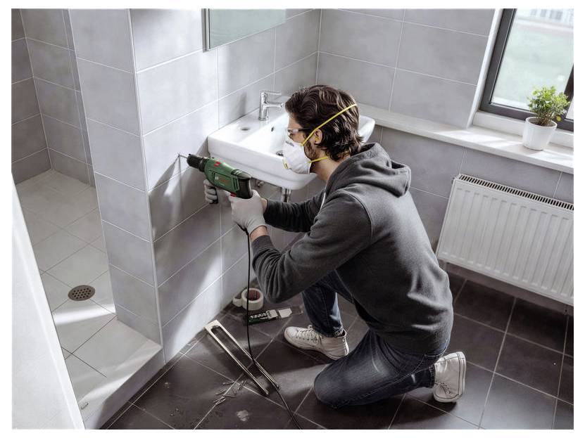 Ein Mann, der eine Maske trägt, bohrt während einer Wohnungsrenovierung mit einem Elektrowerkzeug in eine Badezimmerwand.