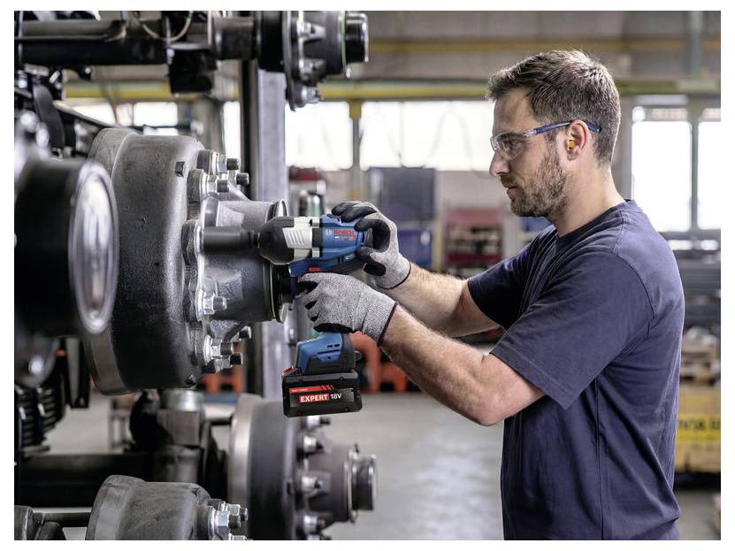 Techniker mit Schutzbrille und Handschuhen benutzt ein kabelloses Elektrowerkzeug, um eine große Komponente einer Industriemaschine in einer Werkstatt festzuziehen.