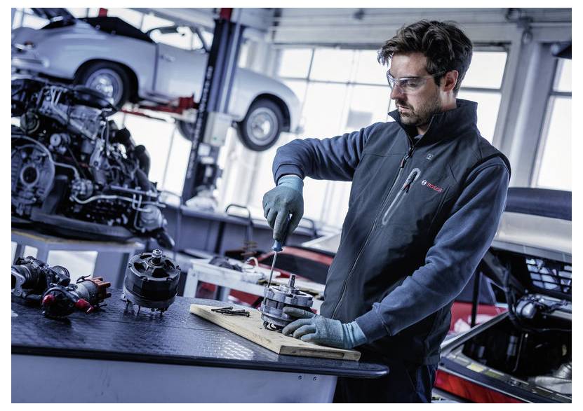 Mechaniker in einer Werkstatt, der Handschuhe und Schutzbrille trägt, benutzt einen Schraubenzieher an einem Autoteil an einer Werkbank, mit Fahrzeugen auf Hubvorrichtungen im Hintergrund.