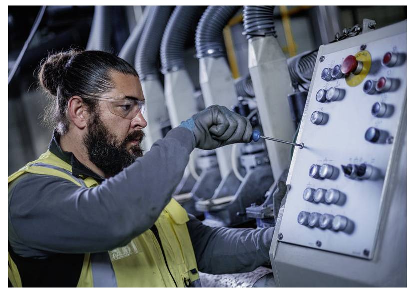 Techniker trägt Schutzbrille und nutzt einen Schraubendreher, um die Steuerungen an einer Bedienungstafel einer Industriemaschine in einer Fabrikumgebung einzustellen.