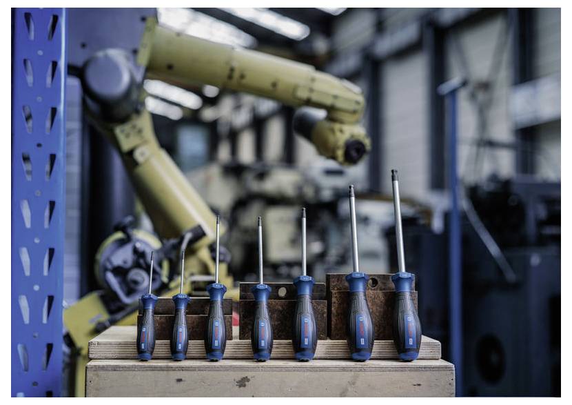 Reihe von Schraubendrehern mit blauen Griffen, die auf einer Holzkiste vor einem industriellen Robotearm in einer Werkstatt angeordnet sind.