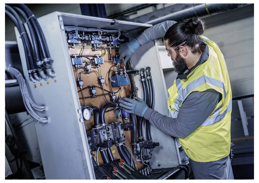 Techniker in einer hochsichtbaren Warnweste inspiziert und justiert die Verkabelung und Schläuche in einem Schrank einer Industriemaschine.