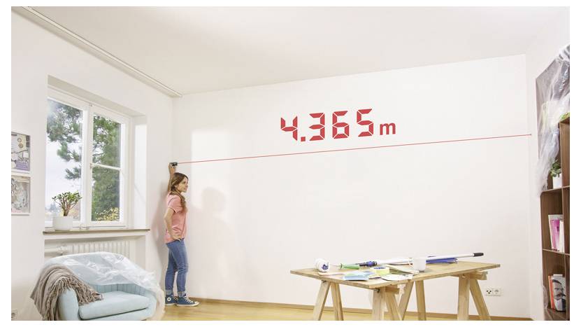 Eine Frau misst die Wandhöhe mit einer Laserwasserwage in einem hellen Raum als „4,365 m", in der Nähe eines Arbeitstisches und eines Fensters.
