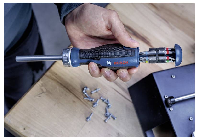 Hand, die einen Bosch-Schraubendreher mit austauschbaren Bits über einer Holzarbeitsfläche hält, mit verstreuten Schrauben in der Nähe.