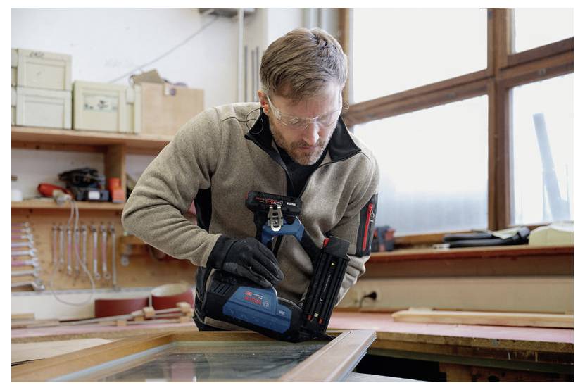 Ein Mann in Schutzbrille verwendet eine kabellose Nagelschussmaschine, um einen Holzrahmen in einer Werkstatt zusammenzusetzen.