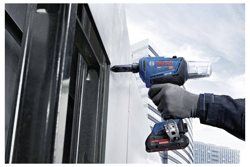 Handschuhgetragener Arbeiter bohrt mit einem blauen Bosch-Elektroschrauber in eine Metallfassade neben einem modernen Gebäude.
