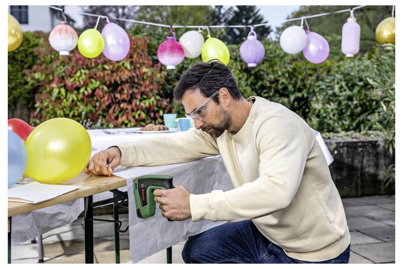Ein Mann benutzt eine tragbare Tacker-Pistole, um ein weißes Tischtuch an einem Tisch bei einer Außenparty-Aufstellung mit farbenfrohen hängenden Laternen und Ballons zu befestigen.