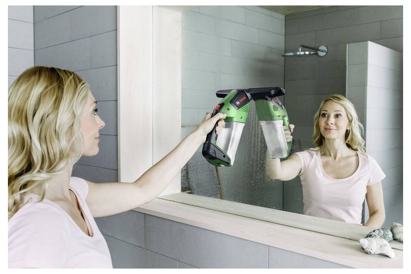 Frau im Badezimmer, die einen Abzieher benutzt, um den Spiegelbehang der Dusche zu reinigen, im Spiegel zu sehen mit einem Duschkopf und Fliesen im Hintergrund.