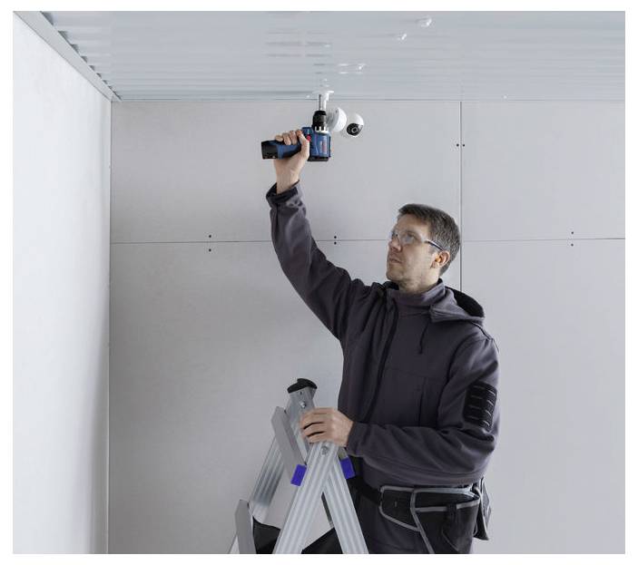 Ein Mann auf einer Stehleiter installiert eine an der Decke montierte Sicherheitskamera in einem Innenraum und benutzt dabei einen Elektroschrauber in einem teilweise fertiggestellten Raum.