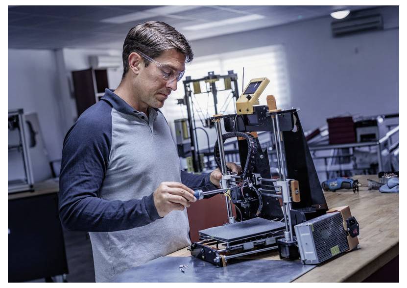 Ein Mann stellt einen 3D-Drucker auf einer Werkbank in einer Werkstatt ein und verwendet ein Werkzeug zum Reparieren oder Kalibrieren der Maschine.