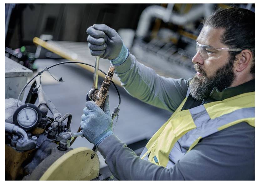 Mechaniker mit Schutzbrille und Warnweste benutzt einen Schraubenschlüssel, um einen Maschinenmotor in einer Werkstatt zu reparieren.