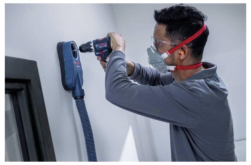 Ein Arbeiter, der eine Schutzbrille und eine Maske trägt, bohrt in eine Wand, während er einen blauen Staubsauger-Staubsammler verwendet, der am Werkzeug befestigt ist.