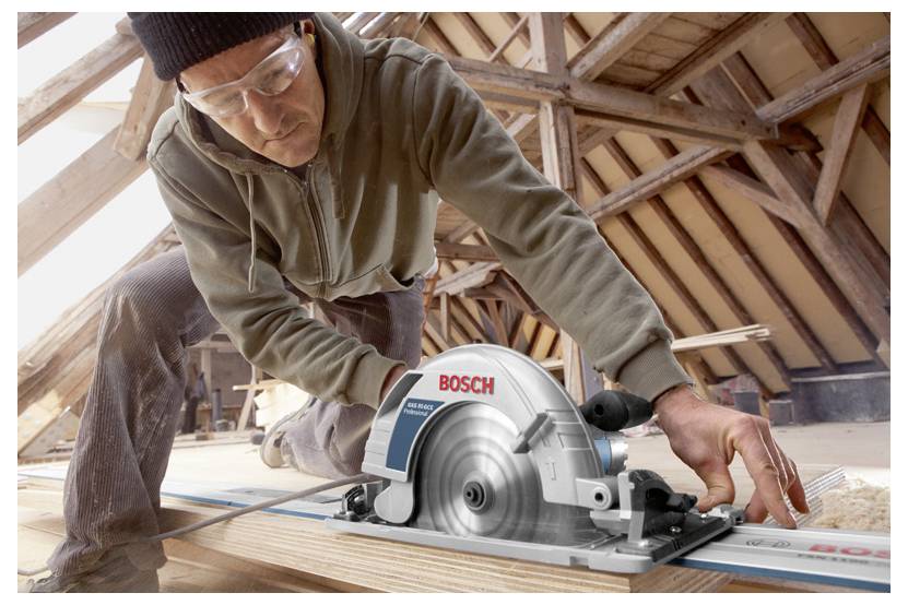 Bauarbeiter, der eine Bosch-Kreissäge verwendet, um ein Holzbrett in einem unfertigen Dachboden mit freiliegenden Dachbalken zu schneiden.