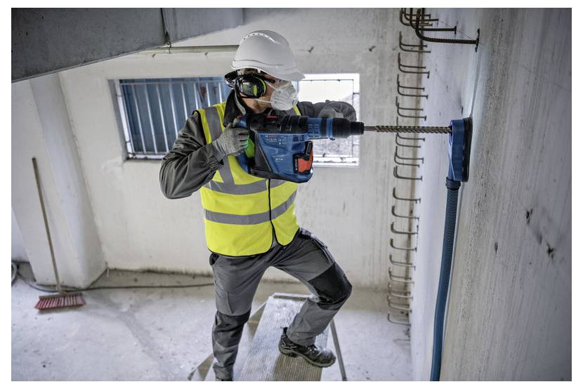 Bauarbeiter mit Schutzhelm und Warnweste, der mit einem Bohrer in eine Betonwand in einem unfertigen Gebäude bohrt.