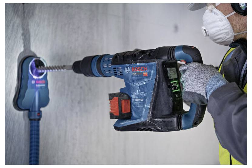Ein Arbeiter bohrt mit einem Bosch-Elektroschrauber ein Loch in eine Betonwand, während er Handschuhe, eine Maske und Schutzausrüstung trägt.