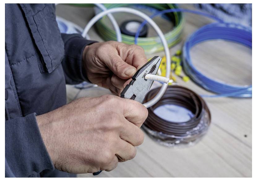 Hände, die eine Zange verwenden, um ein weißes Kabel zu abisolieren, mit Spulen aus farbigem Draht und Kabel im Hintergrund.