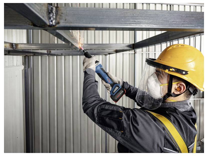 Bauarbeiter mit Helm und Gesichtsschutz verwendet einen Winkelschleiffer zum Schneiden eines Metallträgers, wobei Funken auf einer Baustelle im Innenbereich fliegen.