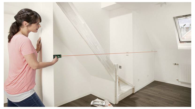 Frau in einem rosa Hemd nutzt eine Wasserwaage mit Laser, um eine Wand in einem leeren Raum mit Treppen und einem Fenster zu vermessen.