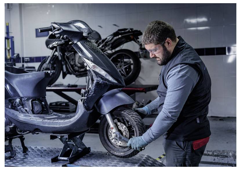 Mechaniker in einer Werkstatt repariert das Vorderrad eines schwarzen Rollers auf einer Hebebühne, mit einem anderen Motorrad im Hintergrund.
