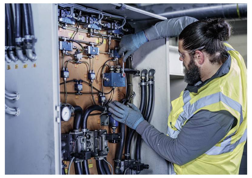 Techniker in einer hochsichtbaren Weste inspiziert und justiert elektrische Steuerleitungen in einem Industrieschaltschrank.