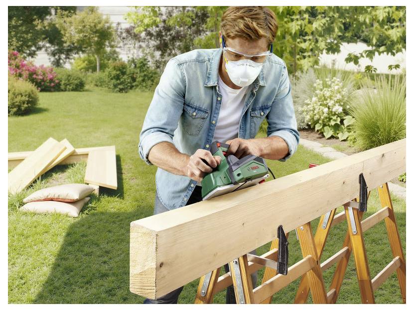 Ein Mann, der eine Maske und eine Schutzbrille trägt, hobelt einen langen Holzbalken auf Böcken in einem Outdoor-Gartenarbeitsbereich.