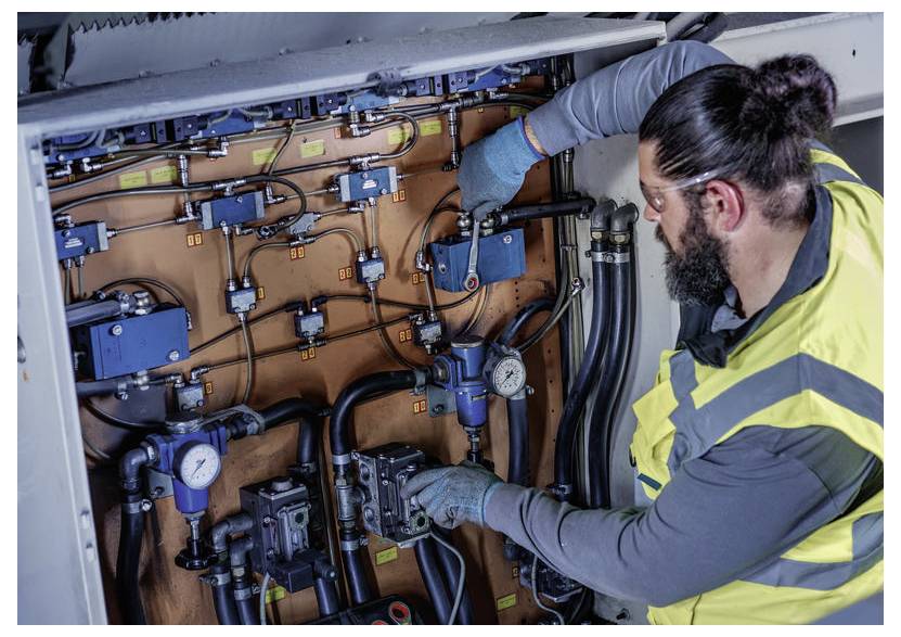 Techniker in Sicherheitsweste und Handschuhen inspiziert und justiert ein Paneel mit Industrierohren, Ventilen, Messgeräten und Steuerboxen in einer Maschine.