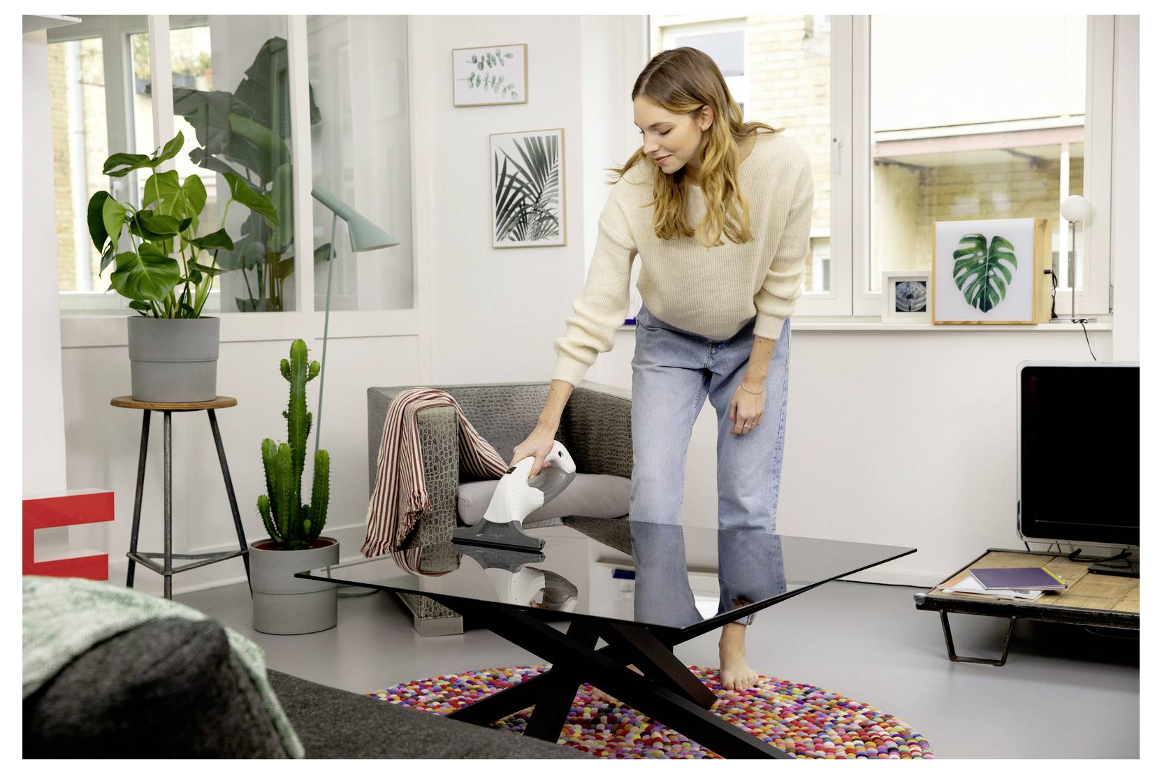 Frau staubsaugt einen gläsernen Couchtisch in einem hellen Wohnzimmer mit Pflanzen und Wandkunst.