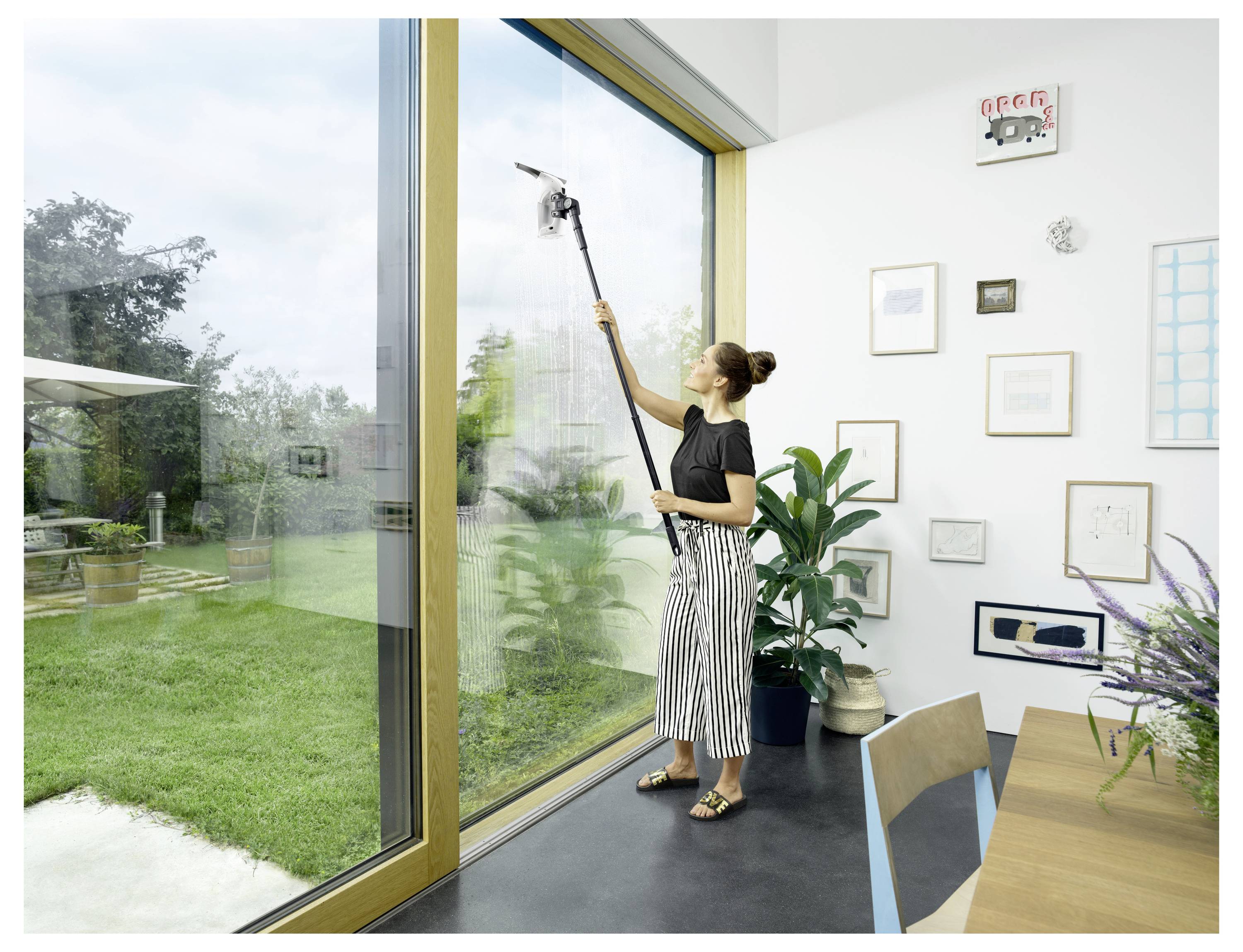 Frau, die einen Fensterabzieher mit langem Griff verwendet, um eine große raumhohe Glastür in einem hellen Raum mit Blick auf einen Garten zu reinigen.