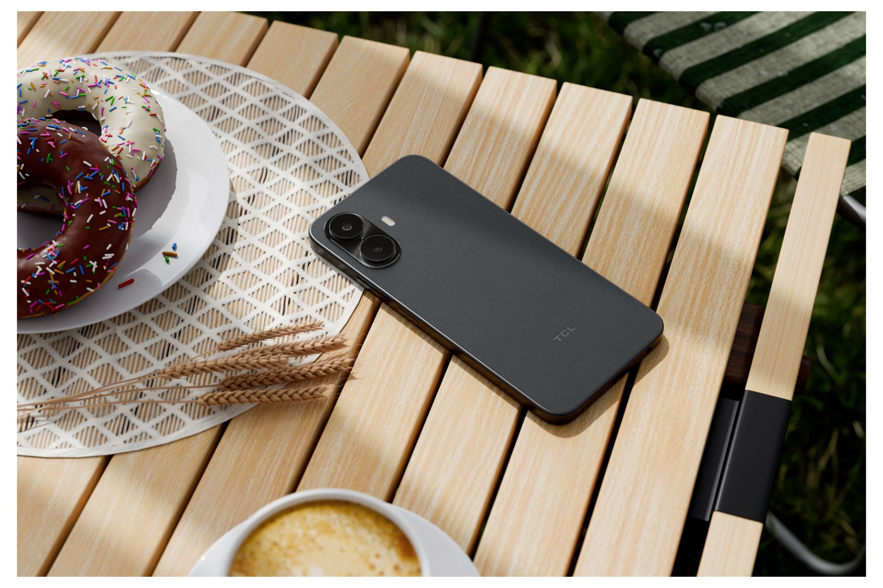 Schwarzes Smartphone auf einem hölzernen Outdoor-Tisch neben zwei Donuts und einer Tasse Kaffee, was eine ungezwungene Frühstücksszene andeutet.