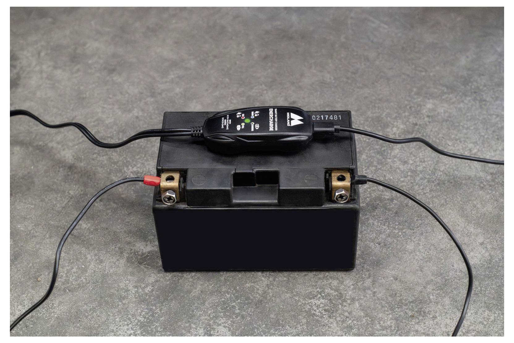 Autobatterie auf Beton mit angeschlossenem Ladegerät oben, mit Ladestatus-Anzeigelichtern und angeschlossenen Kabeln.