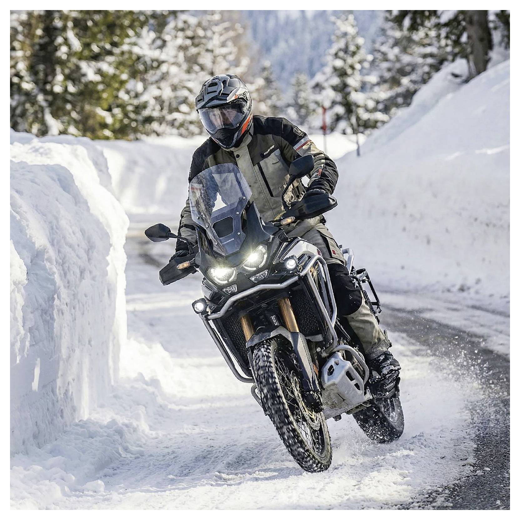 Motorradfahrer auf einem Geländemotorrad auf einem verschneiten Bergpfad fahrend, sich in eine Kurve neigend mit eingeschalteten Scheinwerfern.