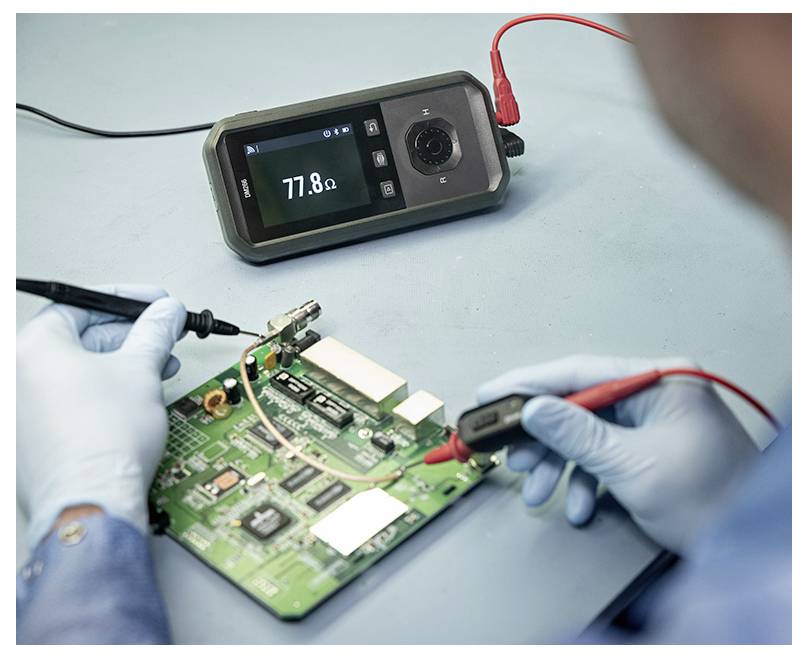 Ein handschuhgetragener Techniker testet eine Leiterplatte mit einem Multimeter; das Messgerät zeigt „77,8Ω" an, während die Messspitzen die Platine berühren.